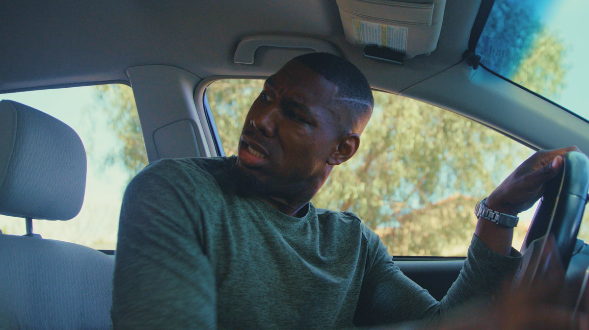A man wearing a dark green long-sleeve shirt sits in the driver's seat of a car, holding the steering wheel and looking to the side.