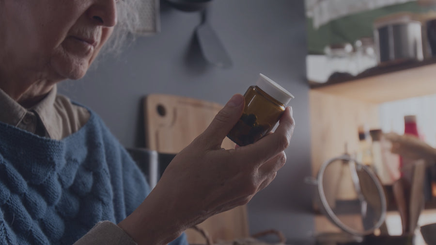 An elderly woman in a blue sweater holds a small amber pill bottle with a white cap in a home setting.