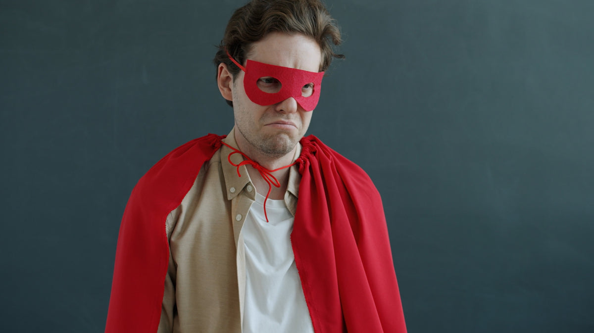A man wearing a red superhero mask and a red cape over a tan shirt and white undershirt, standing against a dark gray background looking sad or upset.