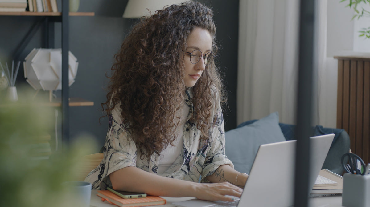 A woman with long curly brown hair and glasses sits at a desk working on a laptop in a modern home office setting.