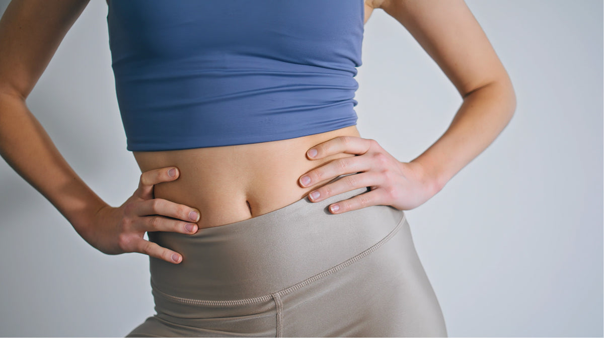 A close-up of a person from the chest down wearing a blue sports bra and beige high-waisted leggings, holding their lower abdomen with both hands against a light gray background.