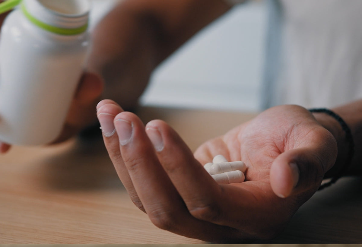 A close-up of a person's hand holding white capsules while another hand pours pills from a white bottle.