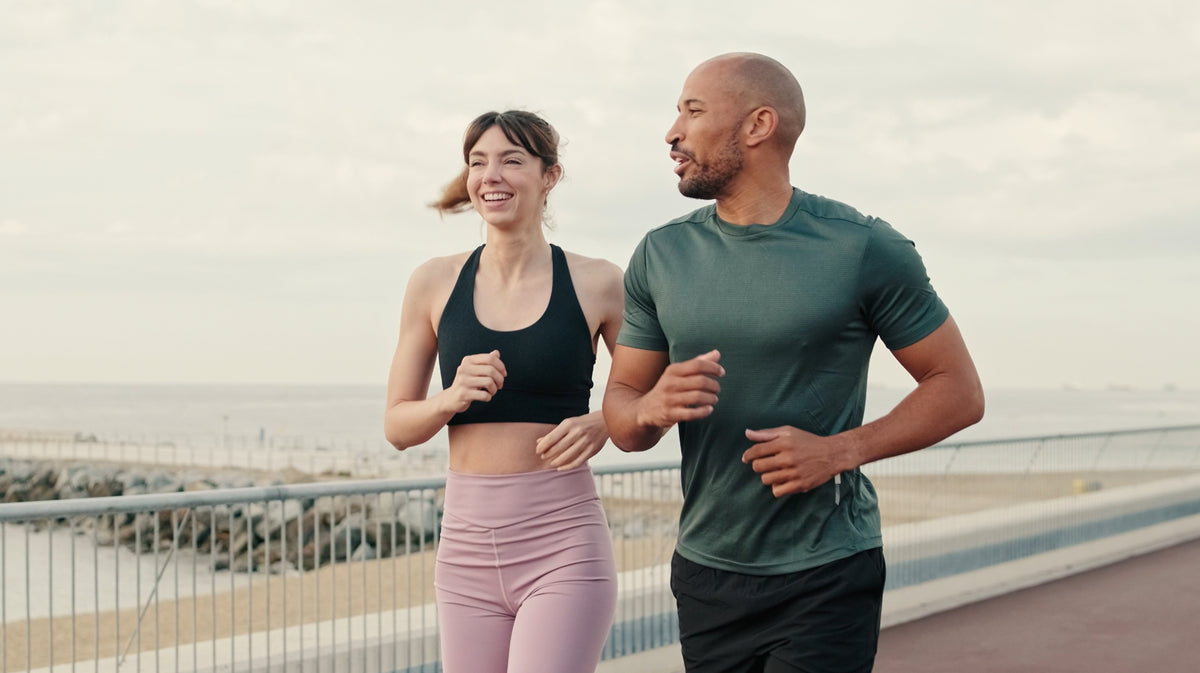 A man and a woman jogging side-by-side on an outdoor coastal path. The woman is wearing a black sports bra and pink leggings and is smiling or entertained by the man, and the man is wearing a dark green t-shirt and black pants talking to the woman.