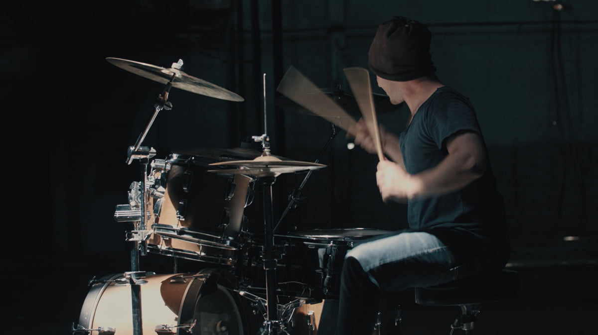 A person wearing a black beanie and dark t-shirt sits behind a drum kit, holding drumsticks and playing in a dimly lit studio setting.