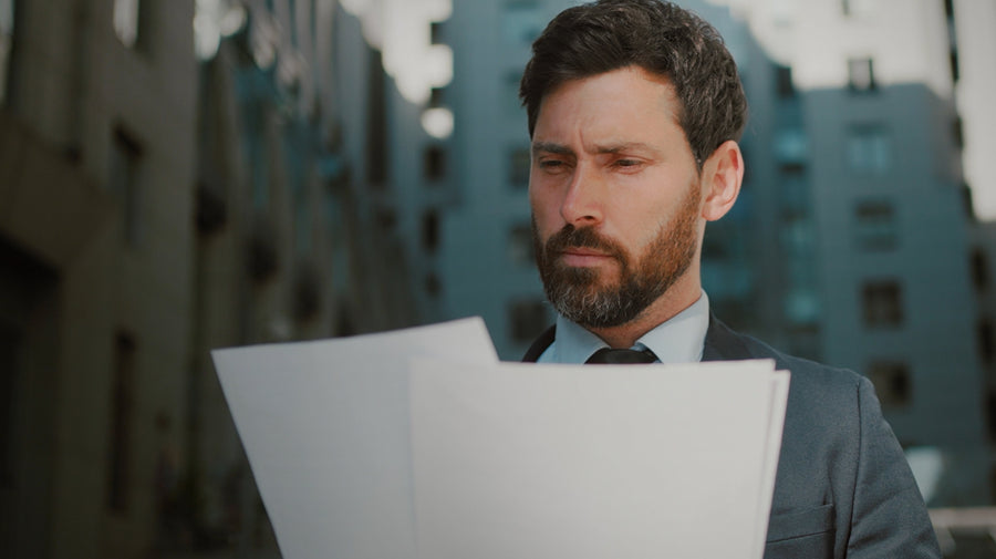 A bearded man in a gray suit, light blue shirt, and dark tie stands outdoors holding several white sheets of paper, with blurred buildings in the background.