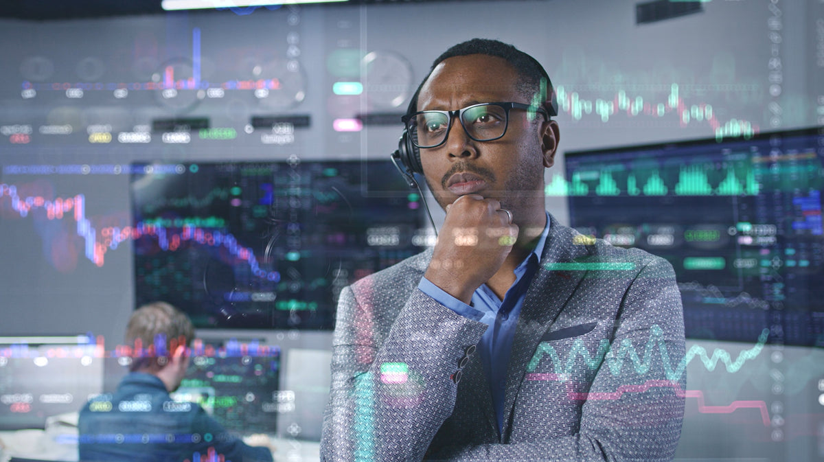 A man wearing glasses, a headset, and a patterned blazer stands in a high-tech trading or control room with glowing charts and graphs visible on screens and overlaid on the image.