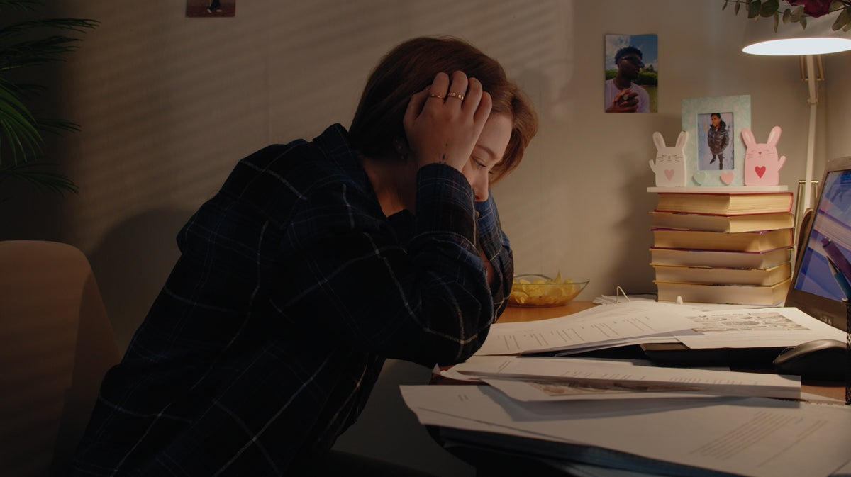 A person sitting at a desk in a dimly lit room, holding their head in their hands, with papers spread across the desk and books stacked on a shelf in the background.