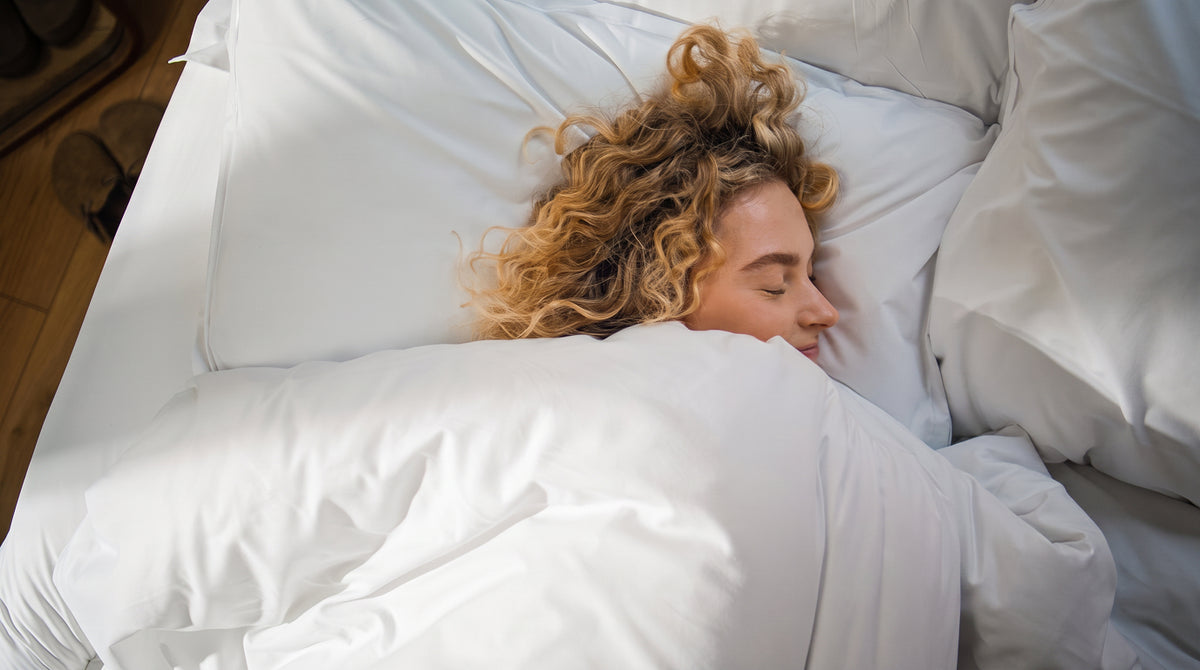 A woman with curly blonde hair sleeping soundly in bed, covered by white pillows and a white duvet.