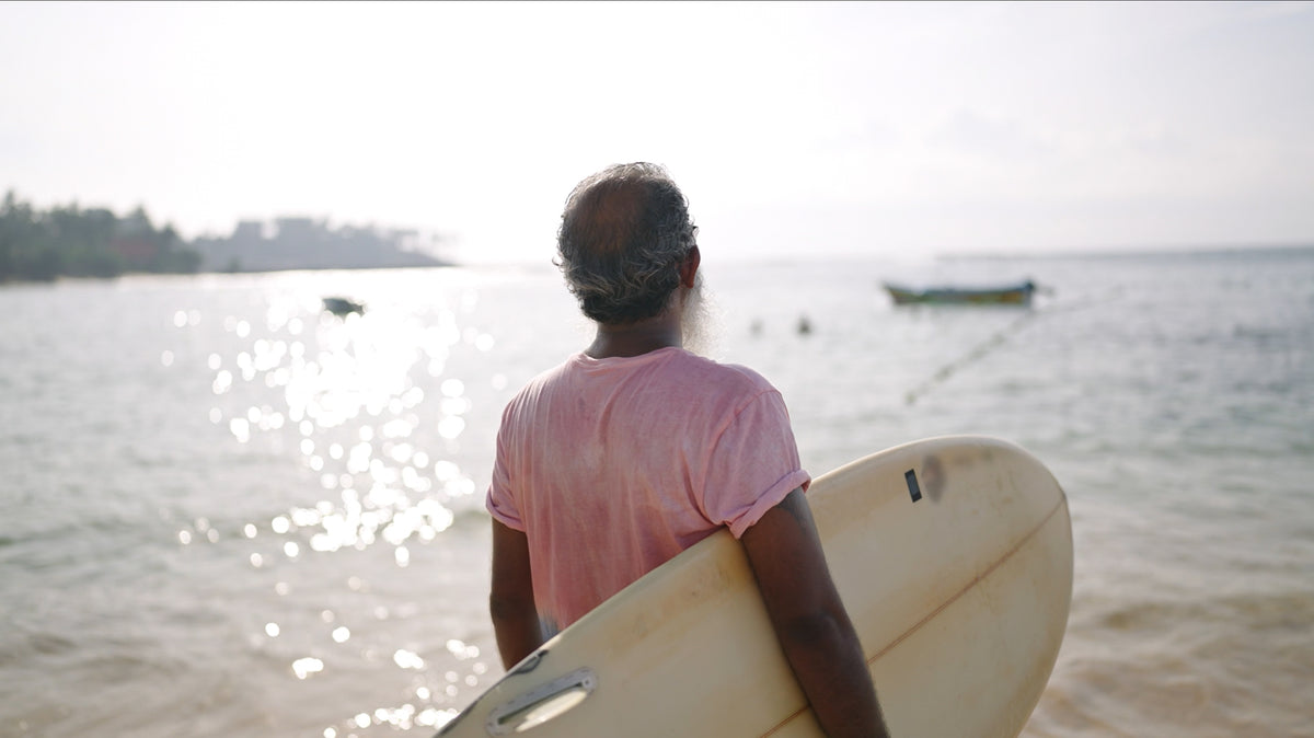 A man seen from behind standing on a beach holding a cream-colored surfboard, wearing a pink t-shirt and looking out towards the ocean.
