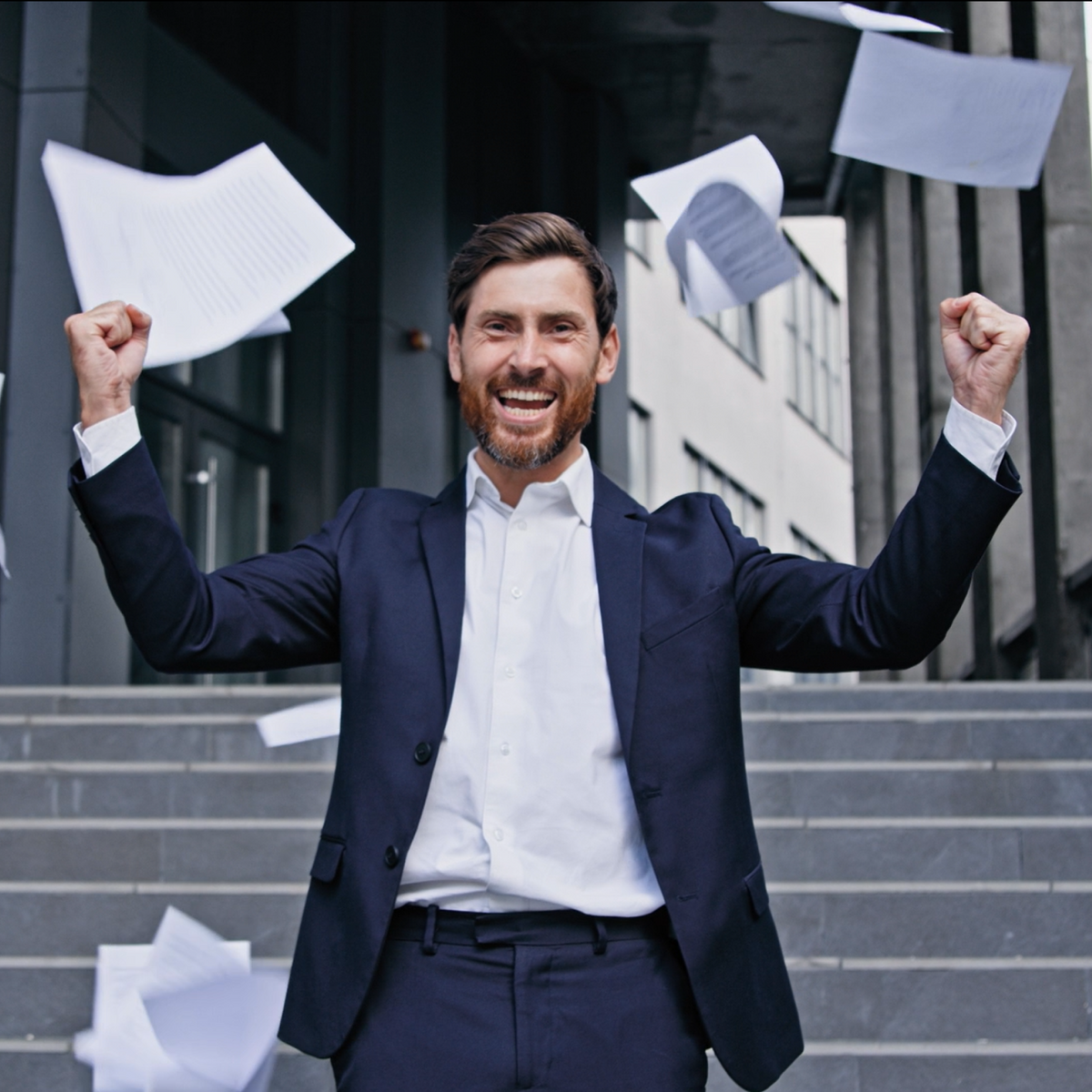 A smiling man in a navy suit and white dress shirt stands on outdoor steps with his arms raised, surrounded by flying papers.