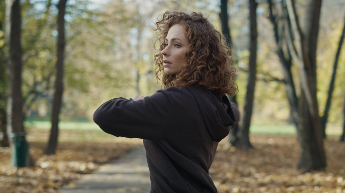 A woman with curly brown hair stretching in a park with autumn trees and fallen leaves.