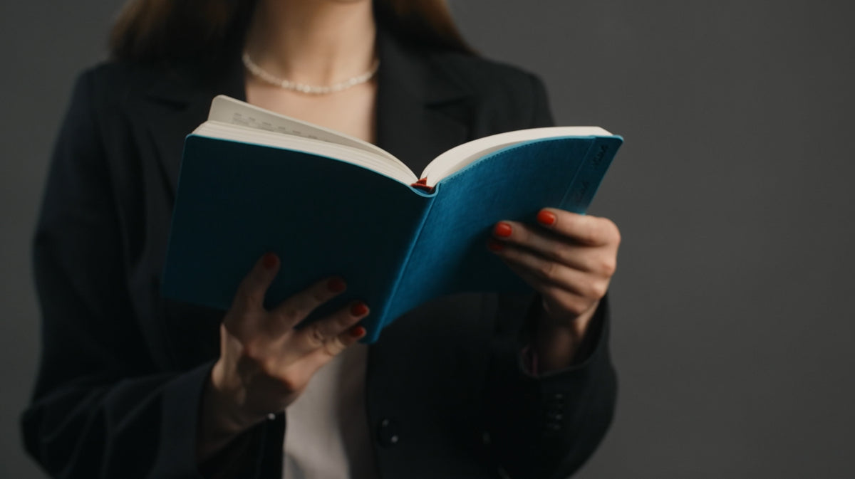 A person in a black blazer holding an open blue notebook or book against a dark gray background.