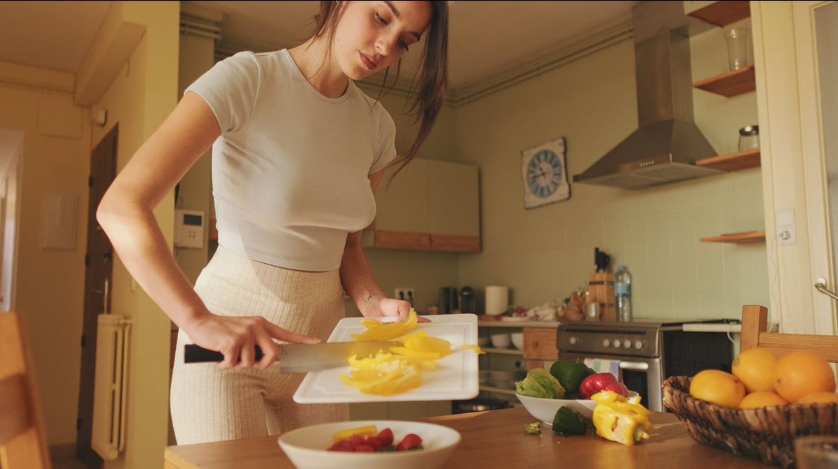 A woman in a white t-shirt and beige pants is standing in a warm, yellow-lit kitchen, slicing yellow bell peppers on a white cutting board.