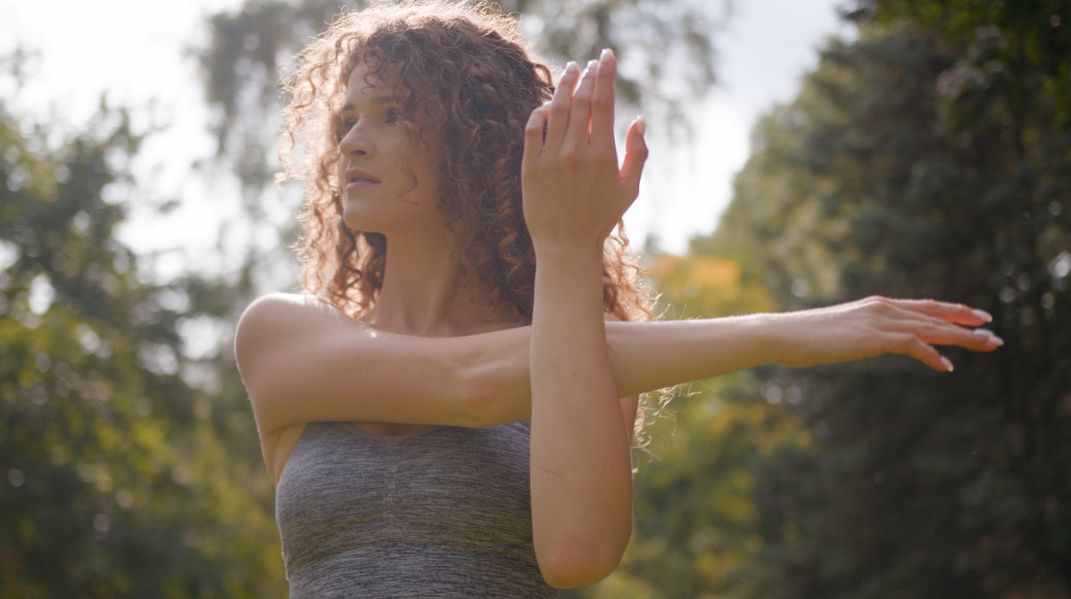A woman with curly hair wearing a gray tank top stretches her arm outdoors in a sunny park.