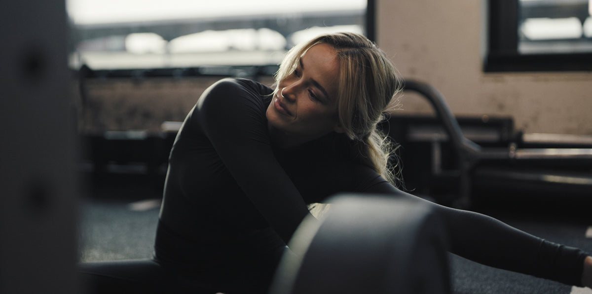 A woman in a black long-sleeve top stretching in a gym setting.