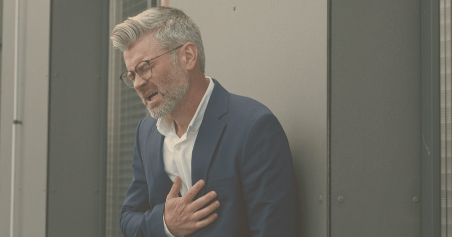 Man in a blue suit and glasses standing against a gray wall holding his chest in discomfort/pain.