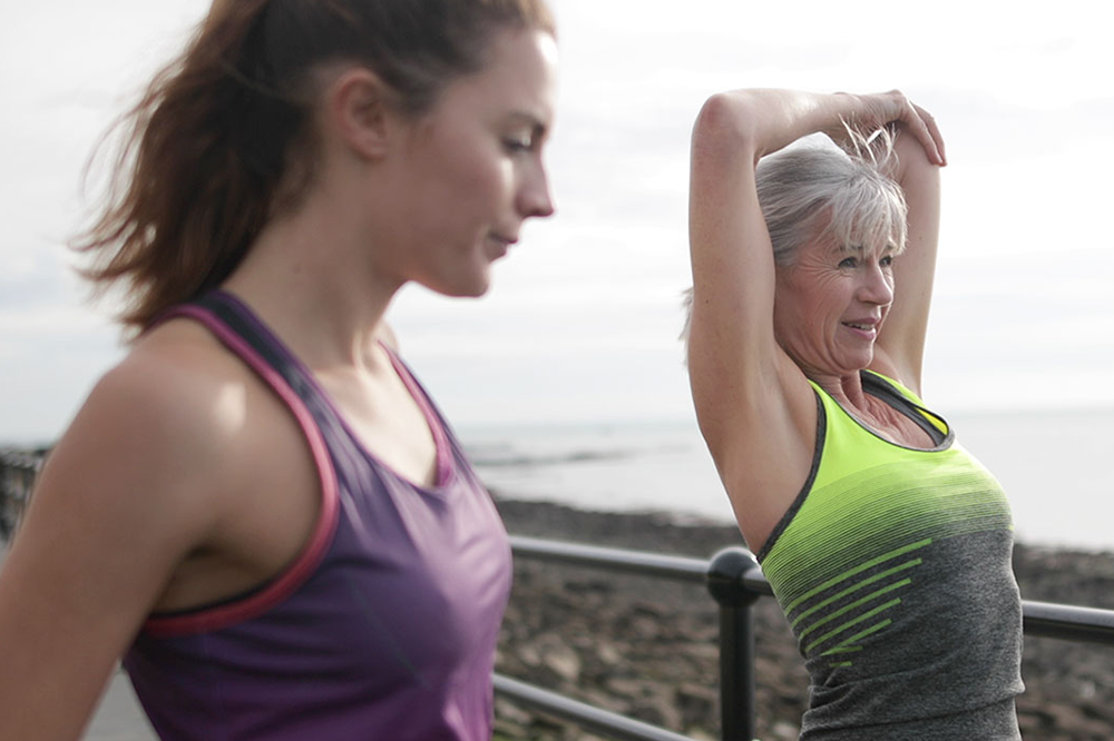 Two women exercising outdoors, one in a purple tank top and the other in a green and gray outfit.