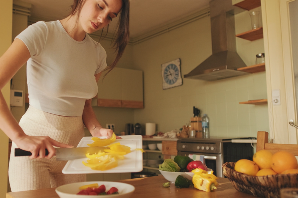 Woman cutting vegetables in a kitchen with various fruits and appliances visible.