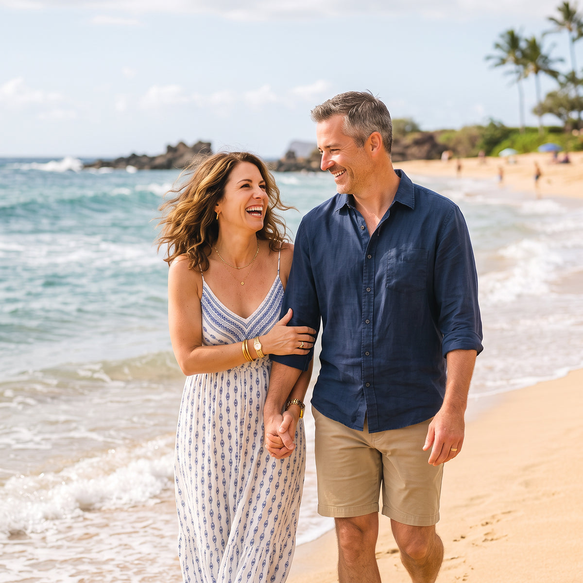 A smiling couple walks arm in arm along a sandy beach with ocean waves in the background. The woman wears a sleeveless white dress with a blue pattern, and the man wears a navy blue button-down shirt and khaki shorts.