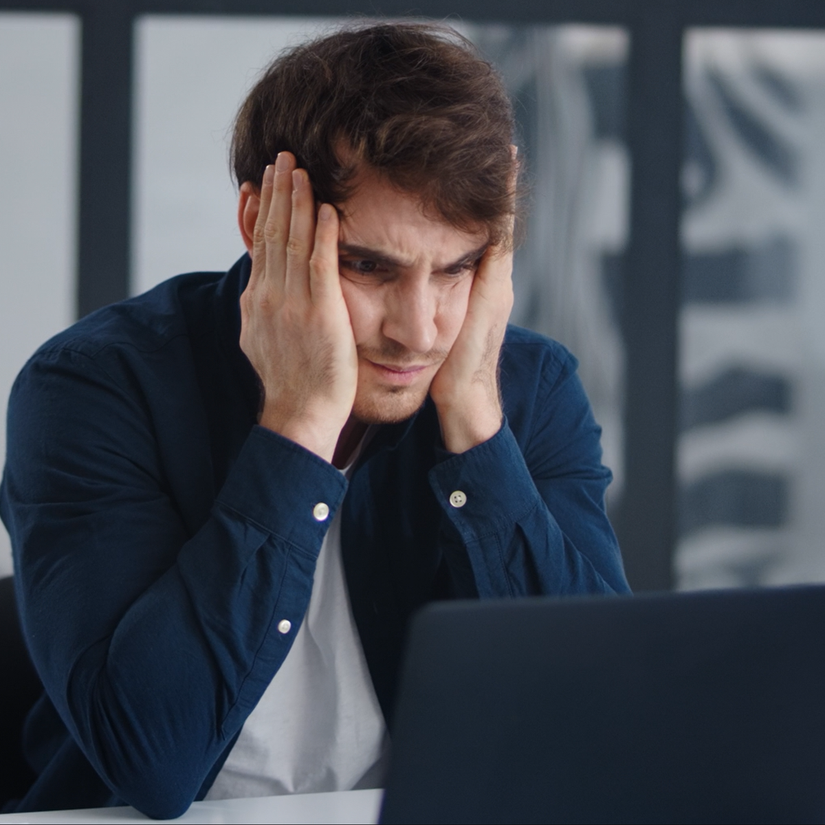 Man in a blue shirt holding his head in front of a laptop, possibly stressed or tired.