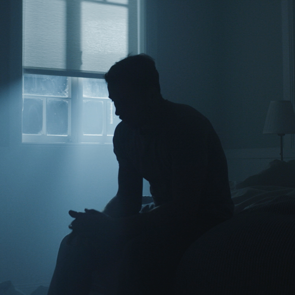 Silhouette of a person sitting by a window with a blue tint in a dimly lit room.
