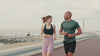 A man and a woman jogging on the boardwalk near water.
