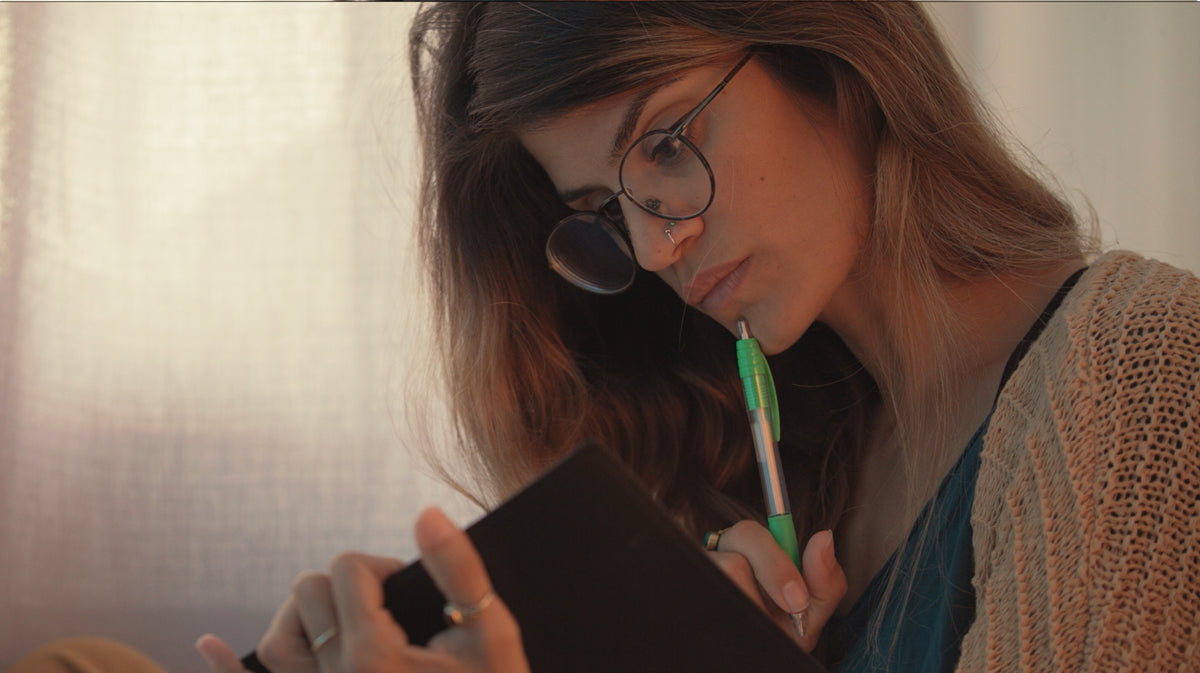 A woman with long brown hair and round black-rimmed glasses holding a green pen and looking down at her notebook studying.