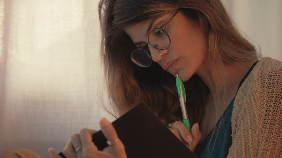 A woman with long brown hair and round black-rimmed glasses holding a green pen and looking down at her notebook studying.